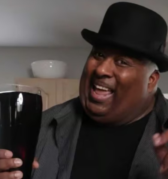 Smiling heavy-set african-american man wearing a
bowler hat, holding a glass boot filled with dark liquid